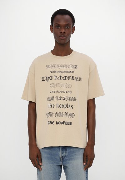 Young man wearing beige oversized T-shirt with "The Kooples" text repeated in various fonts, paired with light blue jeans.