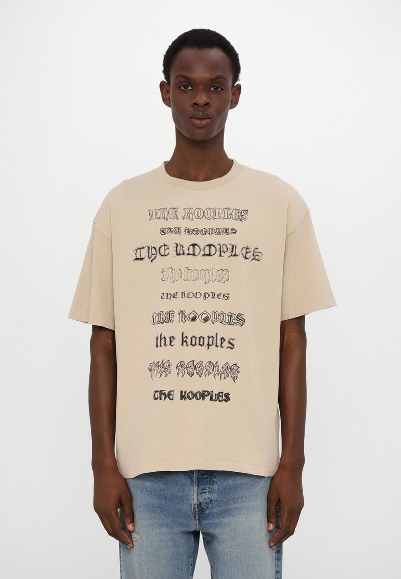 Noor noormees kannab beežite värvi ülekaalulist T-särki, millel on erinevates fontides korduv tekst "The Kooples", koos helesiniste teksadega.