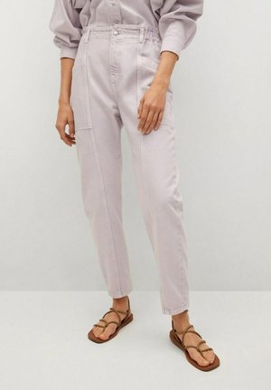Mujer con pantalones de talle alto en lila claro con bolsillos delanteros y sandalias planas marrones con tiras, de pie frente a un fondo liso.