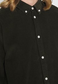 Knapper i mørkegrønt kordfløjlsskjorte krave og øverste bryst med hvide knapper, delvist synlig blont hår.