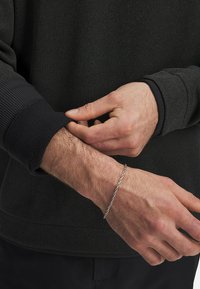 Schwarzer, strukturiert pullover mit gerippten Bündchen. Hand, die den Ärmel anpasst; silbernes, verdrehtesArmband am Handgelenk. Einfaches Design, minimale Details.