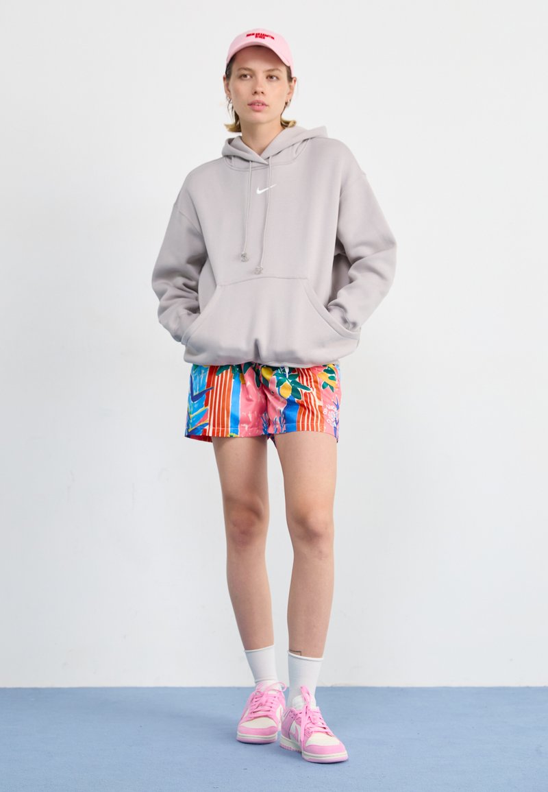 Haut à capuche gris clair avec poche kangourou, shorts fleuris et rayés colorés, chaussettes blanches et baskets roses avec accents blancs.