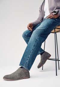 Graue Chukka-Stiefel aus Wildleder mit brauner Sohle, einem minimalistischen Schnürdesign und einer strukturierten Oberfläche, kombiniert mit blauen Jeans.