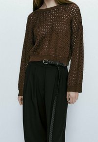 Femme portant un pull à manches longues en tricot ouvert marron, rentré dans un pantalon noir taille haute avec une longue ceinture tressée noire.