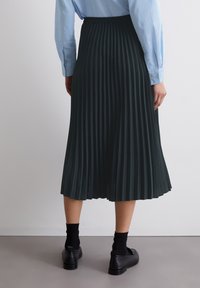Dunkelgrüner plissierter Rock mit elastischem Bund, knielang, aus leichtem Stoff. Kombiniert mit schwarzen Schuhen und Socken.