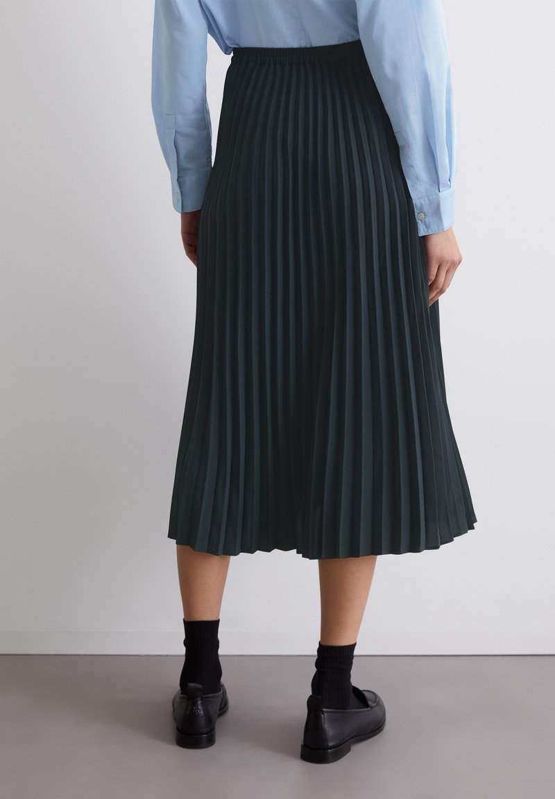 Dunkelgrüner plissierter Rock mit elastischem Bund, knielang, aus leichtem Stoff. Kombiniert mit schwarzen Schuhen und Socken.