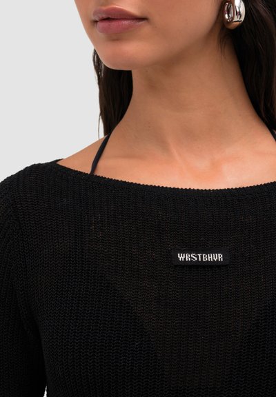 Gros plan d'une personne portant un pull en maille noir avec un patch "WASTEVUR" et une boucle d'oreille argentée en forme d'anneau, montrant le cou et le bas du visage.