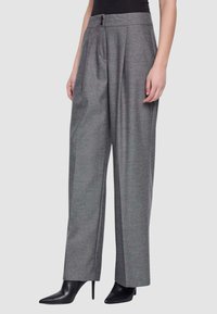 Donna che indossa pantaloni grigio a vita alta e gamba larga abbinati a stivali neri con tacco alto e punta affilata e una maglietta nera.