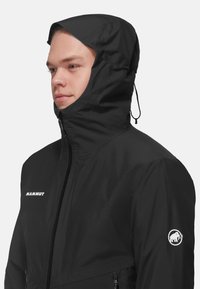 Giacca impermeabile nera con colletto alto e cappuccio regolabile, dotata di un logo sulla manica e tasche laterali con zip.