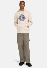 Beige hoodie with a graphic design featuring a figure and text, paired with sage green cargo pants and black shoes.