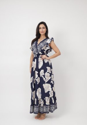 Kobieta w granatowej sukience maxi z białym kwiatowym wzorem i falbankami na rękawach, stojąca z jedną ręką na biodrze na białym tle.