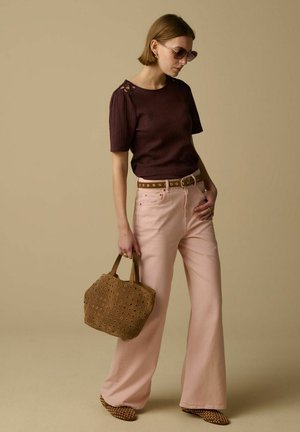 Femme portant des lunettes de soleil, haut marron foncé, pantalon large rose clair, ceinture marron cloutée, tenant un sac à main marron clouté, debout devant un fond beige.