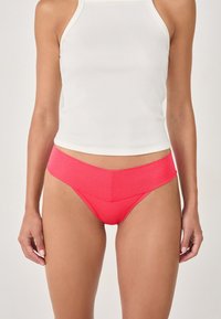 Rote hoch taillierte Bikini-Höschen mit glatter, glänzender Oberfläche und kantigem Design, kombiniert mit einem weißen, figurbetonten Tanktop.
