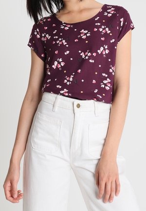 Femme portant un haut à manches courtes violet avec des imprimés floraux roses, rentré dans un pantalon blanc taille haute avec des poches avant.