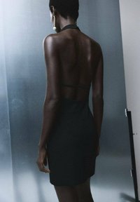 Vestido negro con cuello halter, silueta ajustada, hasta la rodilla con tirantes mínimos, textura suave, que muestra la espalda con exposición de piel.