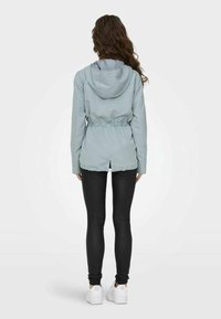 Veste à capuche bleu clair avec taille élastique, cordons de serrage et une fente à l'arrière, associée à des leggings noirs et des baskets blanches.