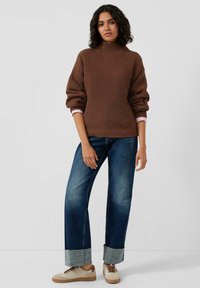 Brązowy sweter z prążkowanego dzianiny z wysokim kołnierzem, połączony z niebieskimi dżinsami o luźnym kroju z podwiniętym mankietem oraz beżowymi trampkami.