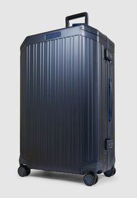 Valigia rigida in blu scuro con scanalature verticali, dettagli blu, maniglia superiore e quattro ruote per la mobilità. Design robusto.