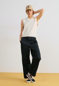 Pulover fără mâneci, tricotat în cabled de culoare crem, asortat cu pantaloni negri largi și adidași negri, completat de o șapcă bej. Fundal uniform.