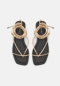 Des sandales plates avec une semelle en cuir noir, ornées de brides métalliques dorées en spirale, créant un style ouvert à l'avant et une lanière autour de la cheville.