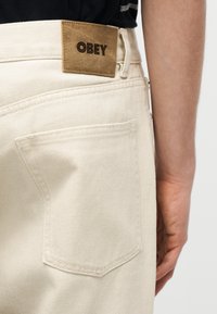 Κοντινή λήψη στην πίσω τσέπη και στην ετικέτα στη μέση μίας μπεζ παντελόνας με το λογότυπο "OBEY", που φοριέται από άτομο με μαύρο πουκάμισο.
