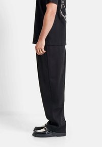 Zwarte oversized t-shirt en zwarte brede broek, gecombineerd met zwarte schoenen met witte accenten en een glanzende afwerking. Eenvoudige achtergrond.