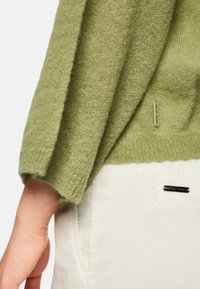 Grüner gestrickter Pullover mit strukturierter Stoffqualität, featuring umgeschlagenen Ärmeln. Kombiniert mit weißen Cordhosen, die ein silbernes Hardware-Etikett tragen.