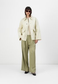 Cream-colored oversized jacket with large front pockets, paired with olive green wide-leg trousers and black high-heel shoes, accessorized with red sunglasses.