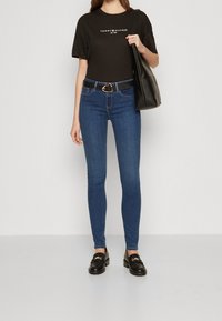 Mujer vistiendo una camiseta negra de Tommy Hilfiger, jeans ajustados azules, cinturón negro, mocasines negros y llevando un gran bolso negro de hombro.