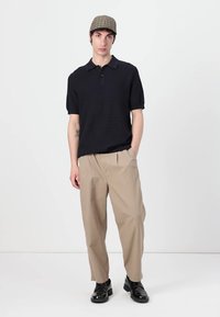 Ung mann iført en rutete flat cap, svart kortermet polo, beige brede bukser og svarte sko, som står med den ene hånden i lommen.