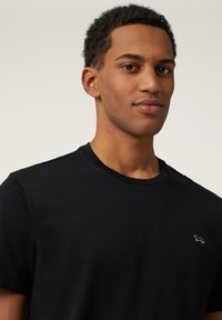 Zwarte katoenen t-shirt met een ronde hals, korte mouwen en een klein grijs logo op de linkerborst. Zacht, gladde textuur. Eenvoudig ontwerp.