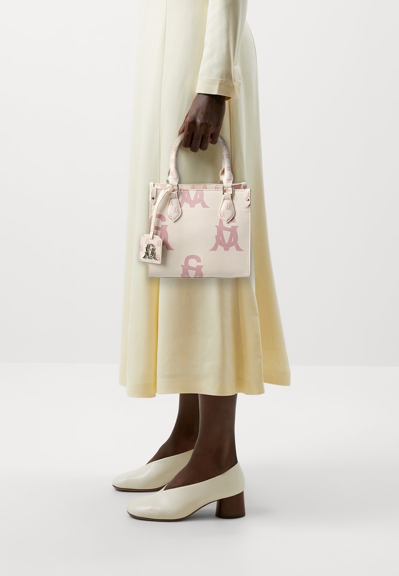 Personne en robe crème tenant un petit sac à main beige avec des lettres roses, portant des chaussures blanches à petit talon, debout sur un fond blanc uni.