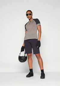 Γκρι ποδηλατικό μπλουζάκι με κοντά μανίκια και μαύρες λεπτομέρειες, σετ με μαύρο σορτς. Η μοντέλο φοράει γυαλιά ηλίου και γάντια, κρατώντας ένα μαύρο κράνος.