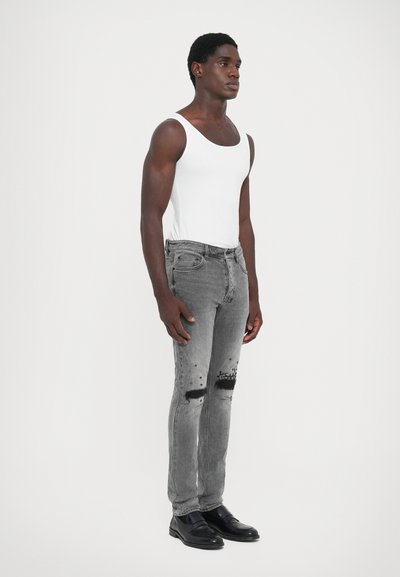 Graue, ausgebleichte Jeans mit Rissen und schwarzen Details an den Knien, kombiniert mit einem weißen Tanktop. Das Model trägt schwarze Schuhe und steht aufrecht.
