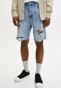 Vaaleansiniset farkkushortsit, joissa on kulutettuja yksityiskohtia ja revittyjä reunoja. Yhdistetty mustiin matalavartisiin lenkkareihin ja harmaisiin ribbikoisiin sukkiin.