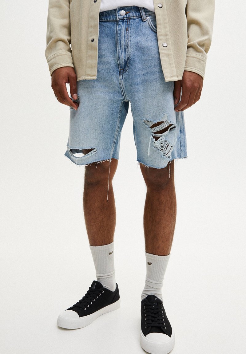 Vaaleansiniset farkkushortsit, joissa on kulutettuja yksityiskohtia ja revittyjä reunoja. Yhdistetty mustiin matalavartisiin lenkkareihin ja harmaisiin ribbikoisiin sukkiin.