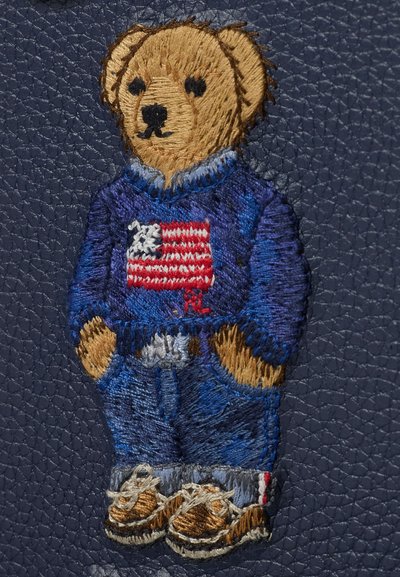 Bordado de oso de peluche marrón con un suéter azul con una bandera estadounidense, jeans azules y zapatos marrones sobre un fondo texturizado azul oscuro.