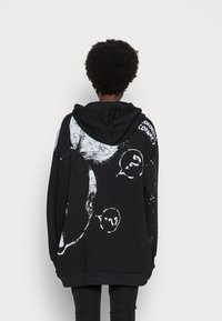 Sudadera oversized negra con un gráfico abstracto blanco en la parte posterior. Cuenta con capucha con cordón y puños acanalados, ofreciendo un ajuste relajado.