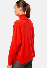 Pull à col roulé rouge en tricot doux et texturé. Coupe oversized avec fentes latérales et poignets côtelés. Porté avec un pantalon sombre.