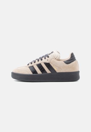 Beige ruskind sneaker med sorte strukturerede striber, sort sål, sorte snørebånd og polstret krave, vist i højre sideprofil.