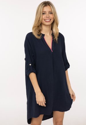 Junge Frau lächelt, trägt ein locker sitzendes dunkelblaues Langarmkleid mit hochgekrempelten Ärmeln und einem pinkfarbenen Saum am Halsausschnitt.