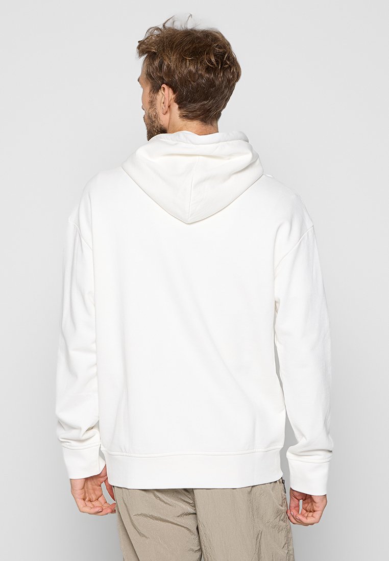 Hombre con cabello castaño claro vestido con una sudadera blanca con capucha y pantalones beige, de pie de espaldas a la cámara contra un fondo gris.