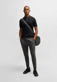 Schwarzes Poloshirt mit gestreiftem Kragen, graue maßgeschneiderte Hose und schwarze Sneakers. Das Model hält eine runde schwarze Tasche und steht vor einem neutralen Hintergrund.