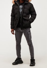 Giacca a vento nera imbottita con cappuccio foderato in pelliccia, abbinata a pantaloni cargo grigi con tasche con zip e sneakers nere con motivi testurizzati.