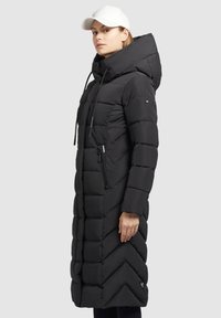 Lang sort pufferjakke med hætte, der har en lynlåslomme på siden og et chevron-stikmønster, lavet af et glat, isoleret materiale.