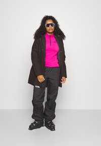 Abrigo impermeable negro, camiseta deportiva de color rosa brillante, pantalones de esquí negros con logotipo y botas de esquí. El modelo lleva grandes gafas de sol blancas. Pelo rizado.
