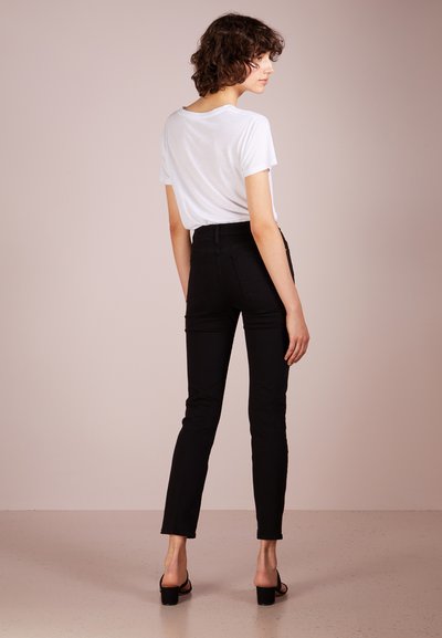 Svarta tajta jeans med hög midja, i kombination med en enkel vit T-shirt. Modellen står med ryggen mot kameran, vilket framhäver den figursydda designen och den stiliga silhuetten.