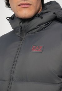 Muškarac nosi tamnosivu jaknu s uključenim jastučićima i visokim ovratnikom, s vidljivim EA7 Emporio Armani logotipom na prsima blizu munje.