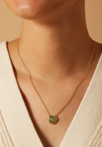 Gouden ketting met een ronde groene edelsteen in een hexagonale setting, gedragen tegen een lichtgekleurde textuurtop.