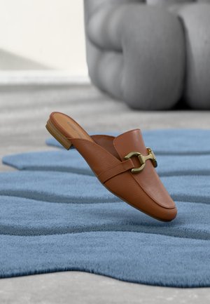 Mocassin en cuir marron à enfiler avec détail mors doré, posé sur un tapis bleu texturé avec un meuble gris coussiné en arrière-plan.